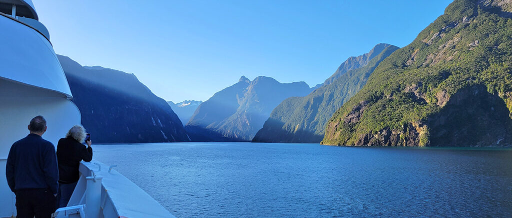 View of a couple on a ship balcony looking ahead as the ship cruises through Milford Sound