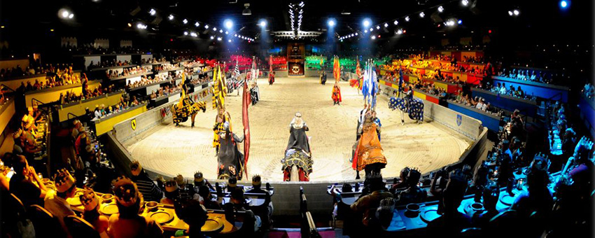 Men and horses dressed in Medieval gear in a ring surrounded by people in stands.