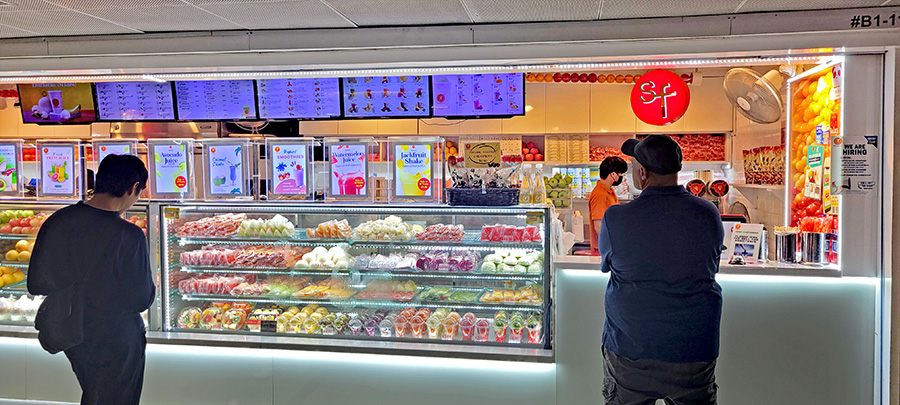Two men stand in front of a counter with fruit and smoothies in an underground shopping area in Singapore