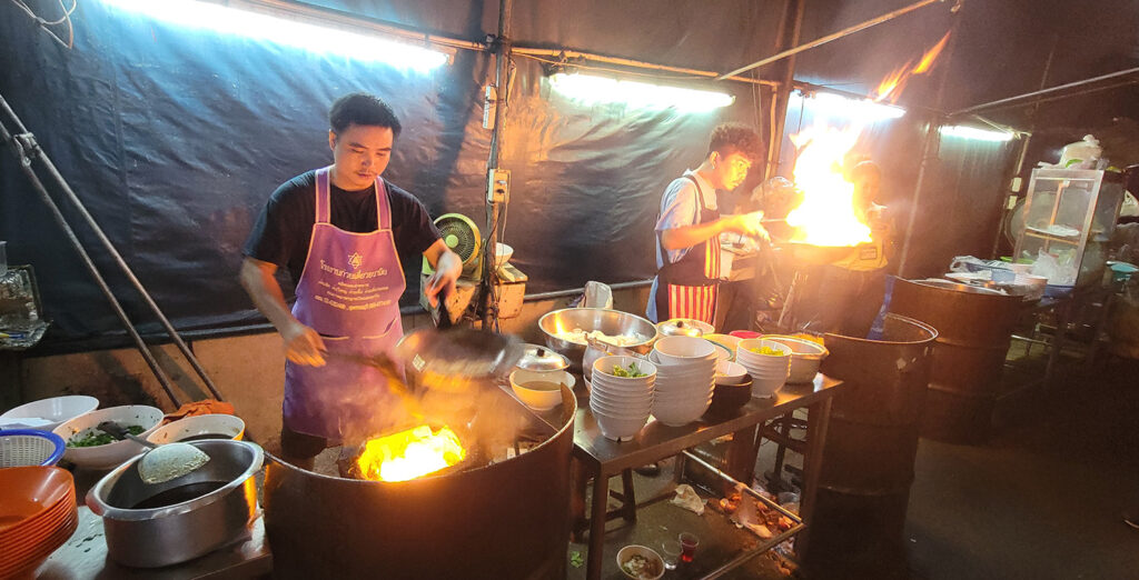 Two men cooking noodles in flaming pots in a back alley kitchen.