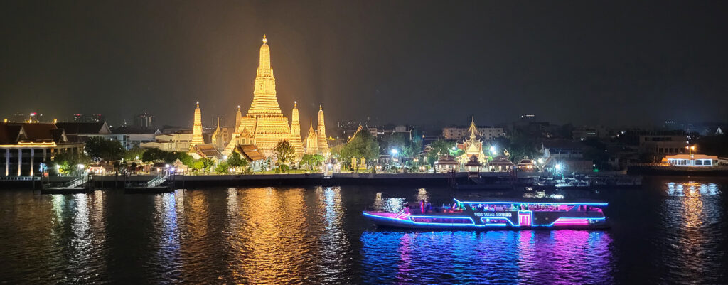 View of a lighted temple and boat passing in front of it. Taken from a rooftop bar in Bangkok.