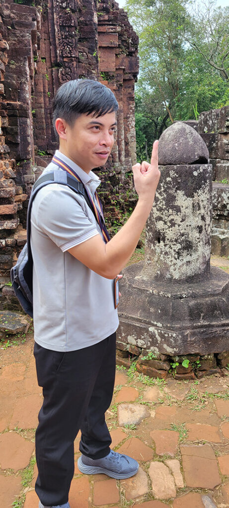 A Vietnamese man holding up a finger with a stone phallic symbol in the background.
