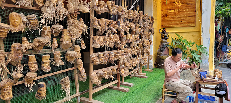 A man carving heads out of bamboo roots with a rack of finished sculptures behind him.