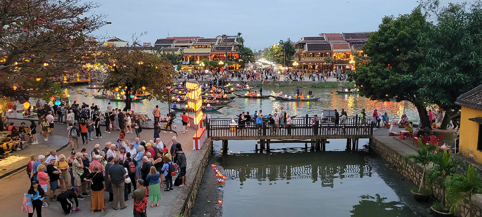 Crowds on a bridge and sidewalks along the canal in Hoi An, Vietnam as night approaches.