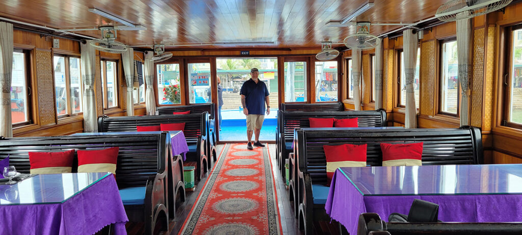 A man stands at the back of a large empty tour boat with tables and wooden seats inside