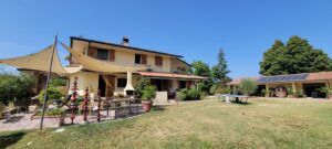 A farmhouse in Italy with a main house and garages separate on the right. There is a long table set up on the back patio covered with a shade cloth.