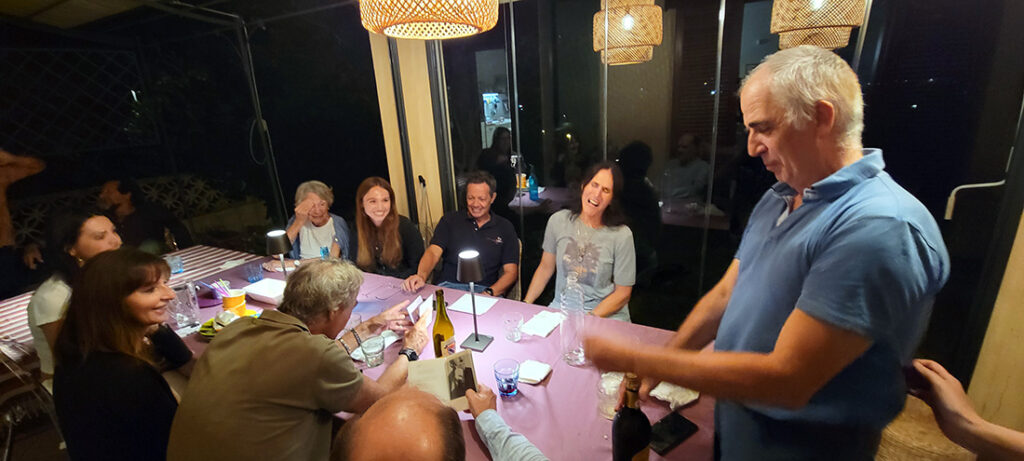 a group of people sitting around a table talking a laughing while a man opens a bottle of proseco