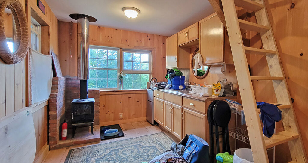 The interior of a cabin with a counter & since, a wood stove, and a ladder that leads upstairs
