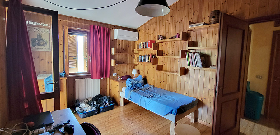 A room with wood paneling and a simple wood single bed. there are shelves over the bed with books and a window next to the bed.