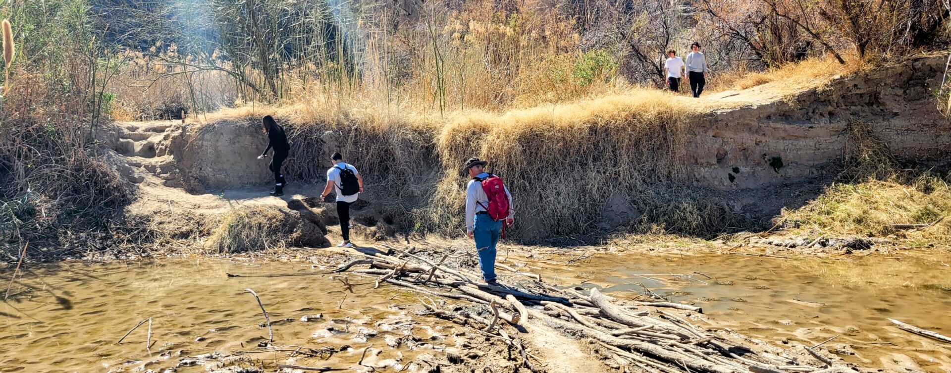 Three people balancing on small logs to cross a small stream. Two additional people are looking down from above.