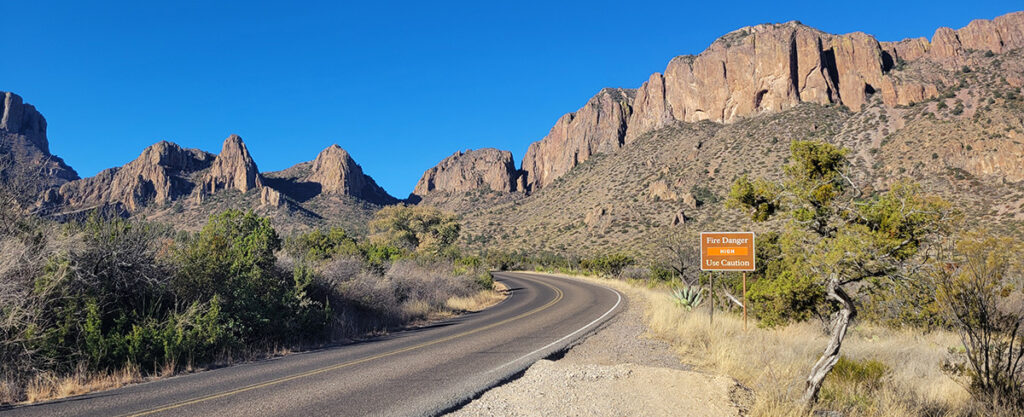 A curved road going through a rocky desert. There is a sign along the road that says the fire danger is "high".