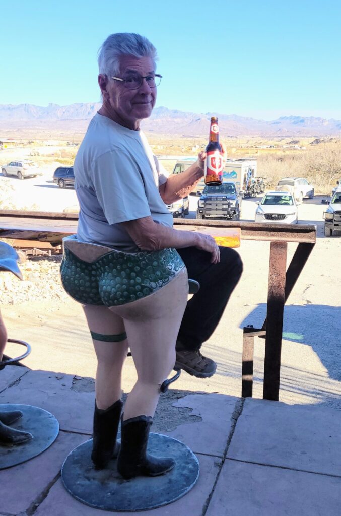 A man sitting on a stool that looks like a large woman in short, shorts. He's holding a beer and the desert is behind him.