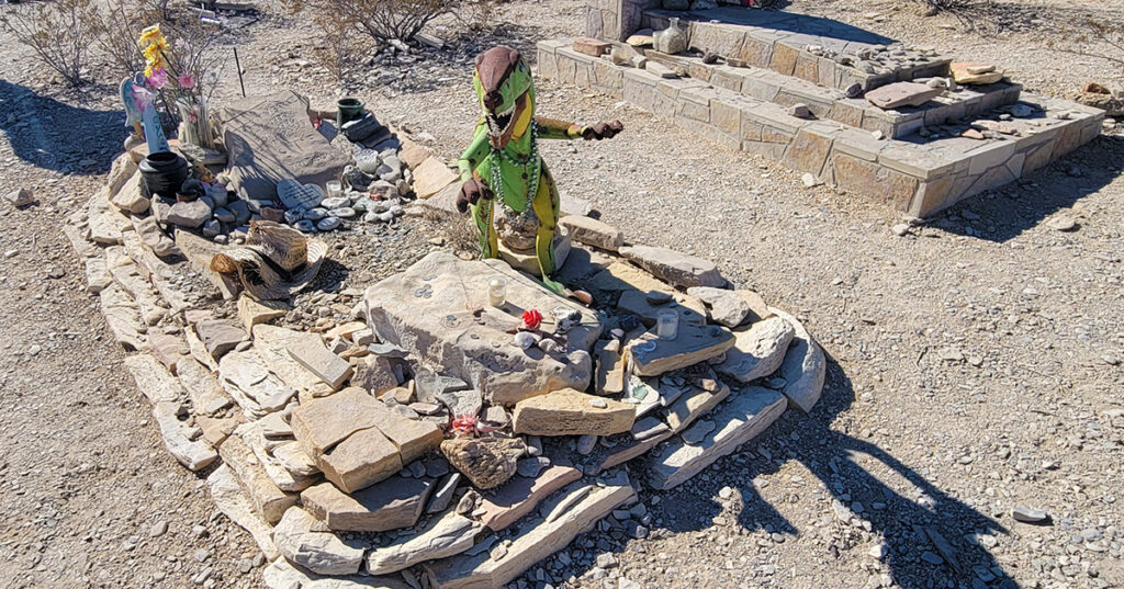 A western grave covered in stones and miscellaneous items including a toy dinosaur.