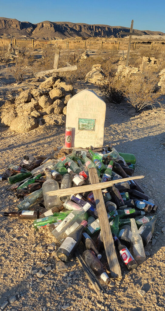 A grave with a head stone and a pile of bottles with a wooden cross on top.