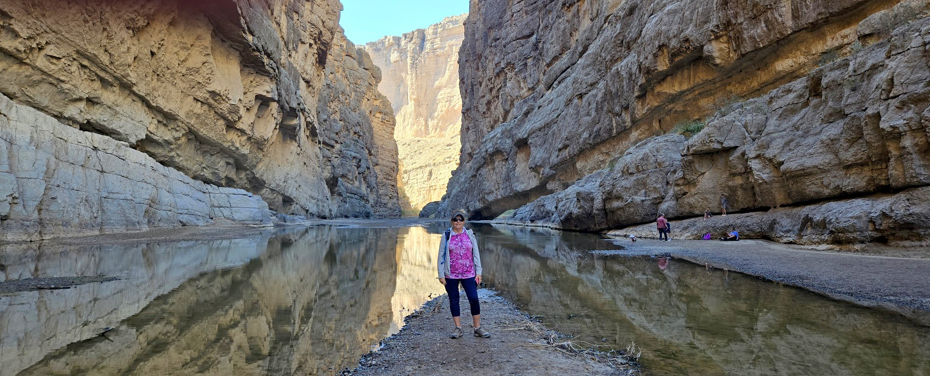 A woman standing on a strip of soil in the middle of a shallow river in a steep canyon.