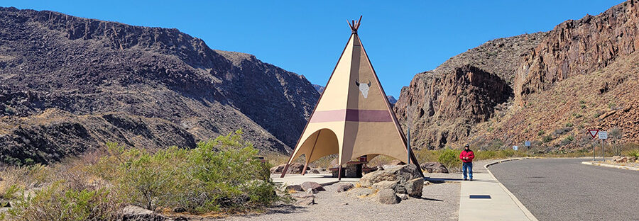 A teepee along a road in the desert with a man wearing a red coat standing next to it